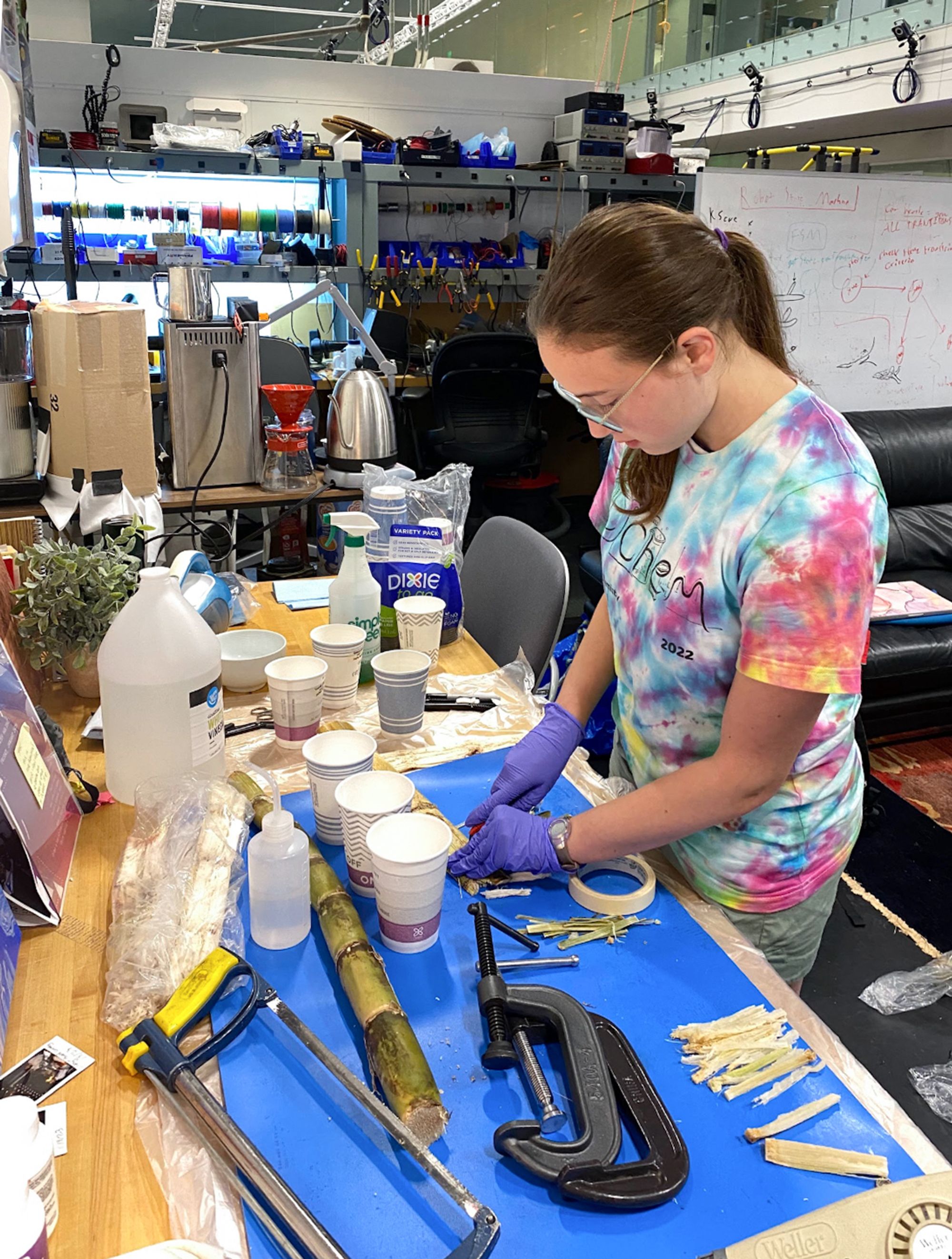 A young woman in safety glasses and gloves works at a lab bench, preparing banana fibers with tools, clamps, and paper cups scattered on the table.